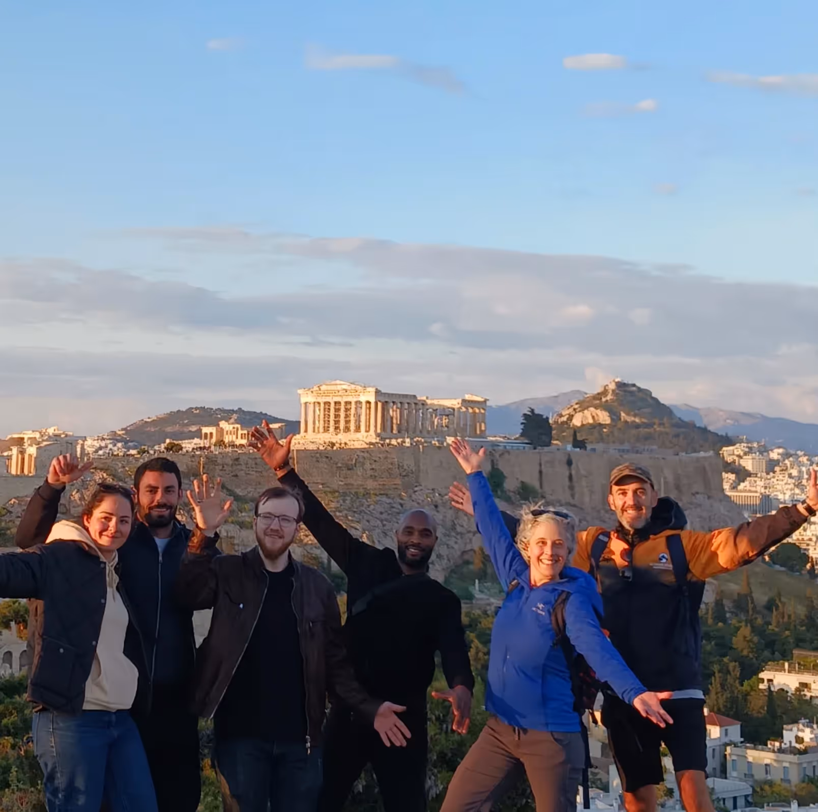 A real Ultimate Athens tour group celebrating with arms raised at the summit, the Acropolis and Lycabettus Hill behind them in the morning light